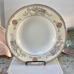 May include: A round, cream-colored plate with a floral design around the rim. The plate is supported by a black metal stand. The floral pattern includes gray and tan flowers and leaves. The plate has a white center.