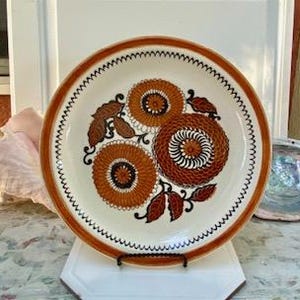 May include: A round, white ceramic plate with a brown rim and a decorative floral design in shades of orange and brown. The plate features a scalloped edge and is displayed on a white stand.