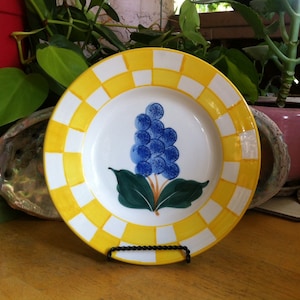 May include: A white ceramic plate with a yellow and white checkered border. The plate features a blue grape cluster design with green leaves. The plate is displayed on a black metal stand, showcasing a decorative and functional piece.