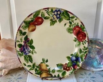 May include: A round, cream-colored ceramic plate with a fruit design. The plate features illustrations of red apples, yellow pears, and purple grapes, surrounded by green leaves. The rim has a decorative border.