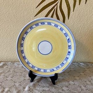 May include: A yellow and white ceramic plate with a blue and white patterned border. The plate is on a black stand, with a beige background and a patterned tablecloth.