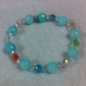 May include: A beaded bracelet featuring alternating turquoise and clear crystal beads, with accents of orange, green, and yellow beads. The bracelet is strung on a clear elastic cord and is suitable for everyday wear.