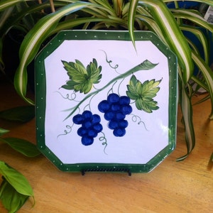 May include: White octagonal serving plate with a green border and a hand-painted design of blue grapes and green leaves. The plate is displayed on a black metal stand, showcasing a classic, decorative style.