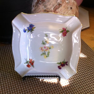 May include: White porcelain square dish with a gold rim and floral designs. The dish features colorful flowers in blue, pink, yellow, orange, and purple. The dish is displayed on a black metal stand.