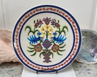 May include: A white ceramic plate with a blue and red border. The plate features a central floral design with blue birds, red flowers, and yellow accents. The plate is displayed on a white stand.