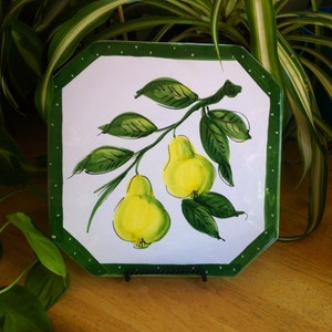May include: Square ceramic plate with a white background and a green border. The plate features a hand-painted design of two yellow pears hanging from a branch with green leaves. The border is a solid green with white polka dots.