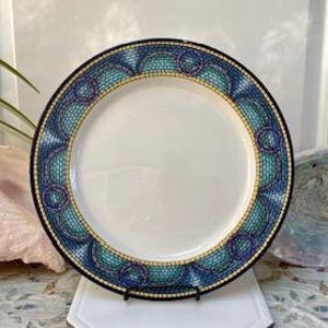 May include: A white ceramic plate with a decorative border featuring a mosaic pattern in shades of blue, teal, and gold. The plate has a black rim and is displayed on a white stand. The plate is approximately 10 inches in diameter.