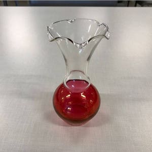 May include: A clear glass vase with a ruffled rim and a round, ruby red base. The vase has a long, slender neck that flares out to the ruffled edge. The base is a deep red color, and the vase appears to be made of glass.