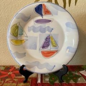 May include: White ceramic plate with a blue rim and a nautical design featuring sailboats with colorful sails. The plate has a diameter of approximately 10 inches and is displayed on a black stand.