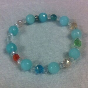 May include: A beaded bracelet featuring turquoise, clear, and colorful beads. The bracelet has large, round turquoise beads interspersed with smaller, clear, faceted beads and a few colorful accent beads. The bracelet is elastic and designed to be worn on the wrist.