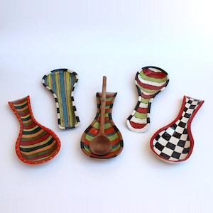 May include: A collection of colorful ceramic spoon rests with unique patterns. One rest holds a wooden spoon. Designs include stripes, checks, and a variety of colors like red, green, black, and yellow. These decorative pieces are for kitchen use.