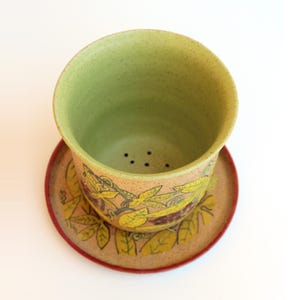 May include: A ceramic tea cup and saucer set. The cup is light green inside and beige on the outside, with a decorative leaf and floral design in yellow and red. The saucer has a red rim and matches the cup's design.
