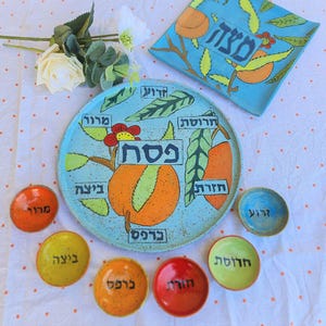 Pode incluir: Uma coleção de pratos e tigelas de cerâmica pintados à mão para o Seder de Páscoa. O prato grande é azul claro com frutas laranja e texto em hebraico. As tigelas menores são de várias cores, cada uma com texto em hebraico.