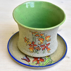 May include: A handmade ceramic planter and saucer set. The planter is beige with a green interior and features a floral design in orange, yellow, and red. The saucer is beige with a blue rim and matching floral accents.