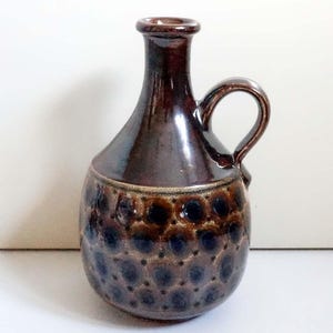 May include: A brown ceramic jug with a handle. The jug features a rounded body with a textured pattern of dark blue and brown circles. The neck and handle are smooth and glossy. The jug is likely vintage.