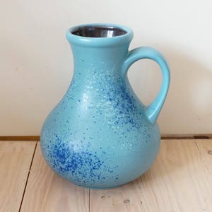Pode incluir: Um vaso de cerâmica azul claro com um esmalte salpicado e uma borda marrom escura. O vaso tem um corpo redondo e uma única alça.