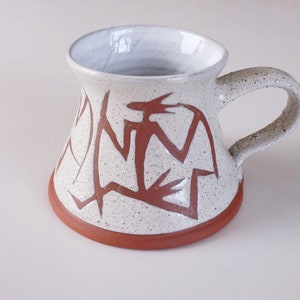 Op de afbeelding: Een witte keramische mok met een bruin handvat en een bruin geometrisch ontwerp. De mok heeft een gespikkeld glazuur en een afgeronde bodem.