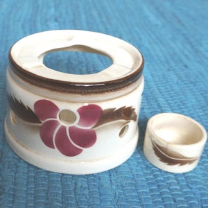 May include: A white ceramic tea warmer with a brown rim and a floral design featuring a pink flower with a yellow centre. The warmer has a small hole in the centre for a teapot.
