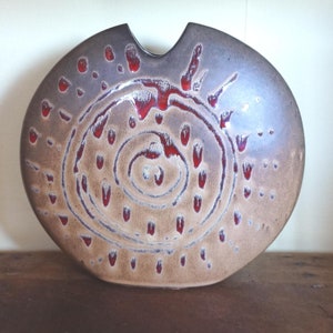 Puede incluir: Un jarrón de cerámica con una forma redondeada única y un corte en la parte superior. El jarrón presenta un diseño en espiral en el centro, con detalles en rojo y una combinación de colores marrón y beige. Es un objeto decorativo.