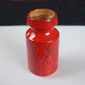 May include: A bright red ceramic vase with a cylindrical body and a slightly flared rim. The vase has etched designs of figures and buildings. The interior is a contrasting tan colour.