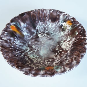 Op de afbeelding: Een bruine keramische kom met witte en oranje glazuur. De kom heeft een golvende, gekrulde rand.