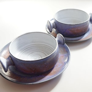 Könnte beinhalten: Zwei Suppenschalen aus Keramik mit Henkeln, jede mit einem weißen Innenraum und einer blauen und violetten Glasur auf der Außenseite. Die Schalen stehen auf passenden Tellern.