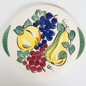 Op de afbeelding: Een witte keramische serveerschaal met groene handvatten heeft een handgeschilderd ontwerp van fruit. Het ontwerp omvat een gele peer, een gele appel en een tros rode druiven. Het fruit wordt omringd door groene bladeren.