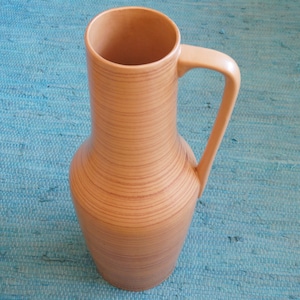 Puede incluir: Un jarr&oacute;n de cer&aacute;mica marr&oacute;n claro, alto, con una boca ancha y un asa. El jarr&oacute;n tiene un patr&oacute;n de veta de madera y est&aacute; sobre una superficie azul.