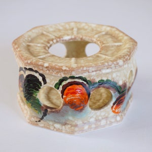 May include: A ceramic tea cosy with a hexagonal shape and a cream-coloured glaze. The cosy has a central hole for a teapot and is decorated with hand-painted floral designs in shades of green, orange, and brown.