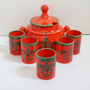 Könnte beinhalten: Ein Set aus sechs orangefarbenen Keramikbechern mit grünen und schwarzen Blumenmustern und einem passenden orangefarbenen Keramik-Krug mit Deckel und Ausguss.