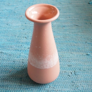 Op de afbeelding: Een kleine, roze keramische vaas met een uitlopende bovenkant en een afgeronde basis. De vaas heeft een witte, getextureerde band rond het midden. De vaas staat op een blauwe geweven mat.