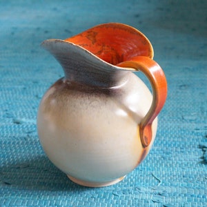 May include: A white ceramic pitcher with a brown glaze and an orange rim. The pitcher has a rounded body and a curved handle.