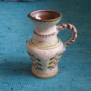 Op de afbeelding: Een witte keramische kan met een bruine rand en een gedraaide handgreep. De kan is versierd met groene en rode bloemenmotieven.