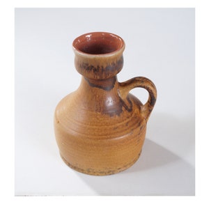 Op de afbeelding: Een kleine, bruine keramische vaas met een handvat en een bruine glazuur. De vaas heeft een licht uitlopende bovenkant en een rond lichaam.