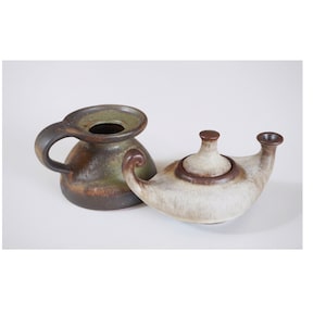 Op de afbeelding: Twee keramische olielampen, een met een handvat en een brede opening, de andere met een tuit en een deksel. De lamp met het handvat is bruin en groen, terwijl de andere lamp wit is met bruine accenten.