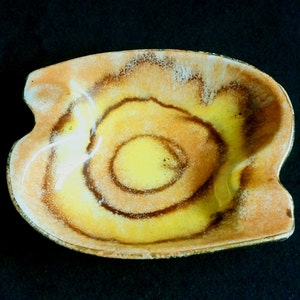 May include: A yellow and brown ceramic ashtray with a swirl design. The ashtray is shaped like a leaf and has a slightly raised edge.