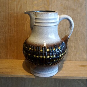 May include: A white ceramic jug with a brown and yellow geometric pattern. The jug has a wide base and a narrow neck with a spout.