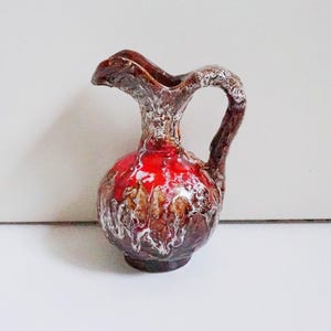 Può includere: Un piccolo brocca decorativa in ceramica con corpo bombato e manico. La brocca presenta una vivace smaltatura rossa che sfuma in marrone e bianco, creando un aspetto unico e strutturato. Il manico e il beccuccio sono di un marrone più scuro.
