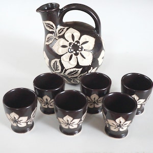 Op de afbeelding: Een set van bruine keramische bekers en een kan met witte bloemenmotieven. De kan heeft een handvat en een rond lichaam. De bekers zijn klein en hebben een ronde vorm.