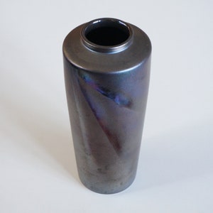 May include: A dark grey ceramic vase with an iridescent glaze. The vase has a cylindrical shape and a smooth, glossy finish.