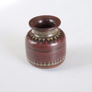 Op de afbeelding: Een kleine, bruine en crèmekleurige keramische vaas met een geometrisch patroon. De vaas heeft een rond lichaam en een licht uitlopende opening.