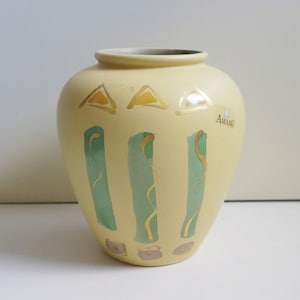 Peut inclure: Un vase en céramique crème avec un corps arrondi et un col étroit. Le vase est décoré de triangles dorés, de rayures verticales vertes et de volutes dorées. Le mot "AMANO" est imprimé sur le côté.