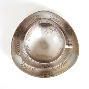 Op de afbeelding: Een vintage theekopje, schotel en bordenset. De set heeft een lichtgrijze achtergrond met gouden filigraan en bloemmotieven. De kop en schotel zijn rond, en het bord is driehoekig.