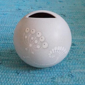 以下が含まれることがあります： 上部に開口部のある丸いオフホワイトのセラミック花瓶。花瓶は繊細な白い花の模様で装飾されています。背景は質感のある青色の表面です。
