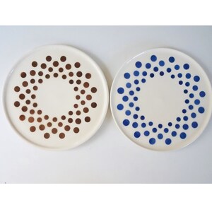 Op de afbeelding: Twee witte keramische borden met een cirkelvormig patroon van stippen. Het ene bord heeft gouden stippen en het andere heeft blauwe stippen.