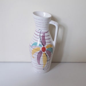 Op de afbeelding: Witte keramische vaas met een bloemenmotief in paars, geel, rood en blauw. De vaas heeft een handvat en een gestreept patroon.
