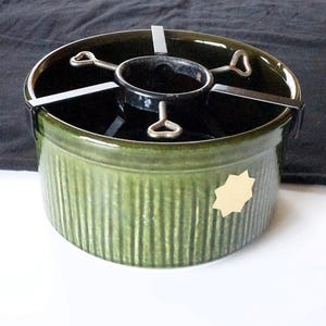 Puede incluir: Un soporte de cerámica verde para árbol de Navidad con un soporte de árbol de metal negro. El soporte tiene detalles acanalados verticales y una pequeña pegatina en forma de estrella en blanco en el lateral. El soporte está diseñado para sostener un árbol de Navidad.