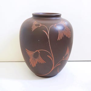 Op de afbeelding: Een bruine keramische vaas met een afgeronde body en een smalle hals. De vaas is versierd met een oranje bloemenontwerp. Het bloemenontwerp toont gestileerde bloemen en stelen. De vaas is een decoratief item.