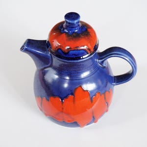Op de afbeelding: Een blauwe theepot van keramiek met een deksel en een handvat. De theepot heeft een rood en oranje glazuur met een druppel effect.
