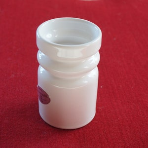 Op de afbeelding: Een witte keramische kandelaar met een cilindrische basis en een uitlopende bovenkant. De kandelaar heeft een geribbeld ontwerp in het midden en een kleine, roze sticker aan de zijkant. Het object staat op een rode ondergrond.